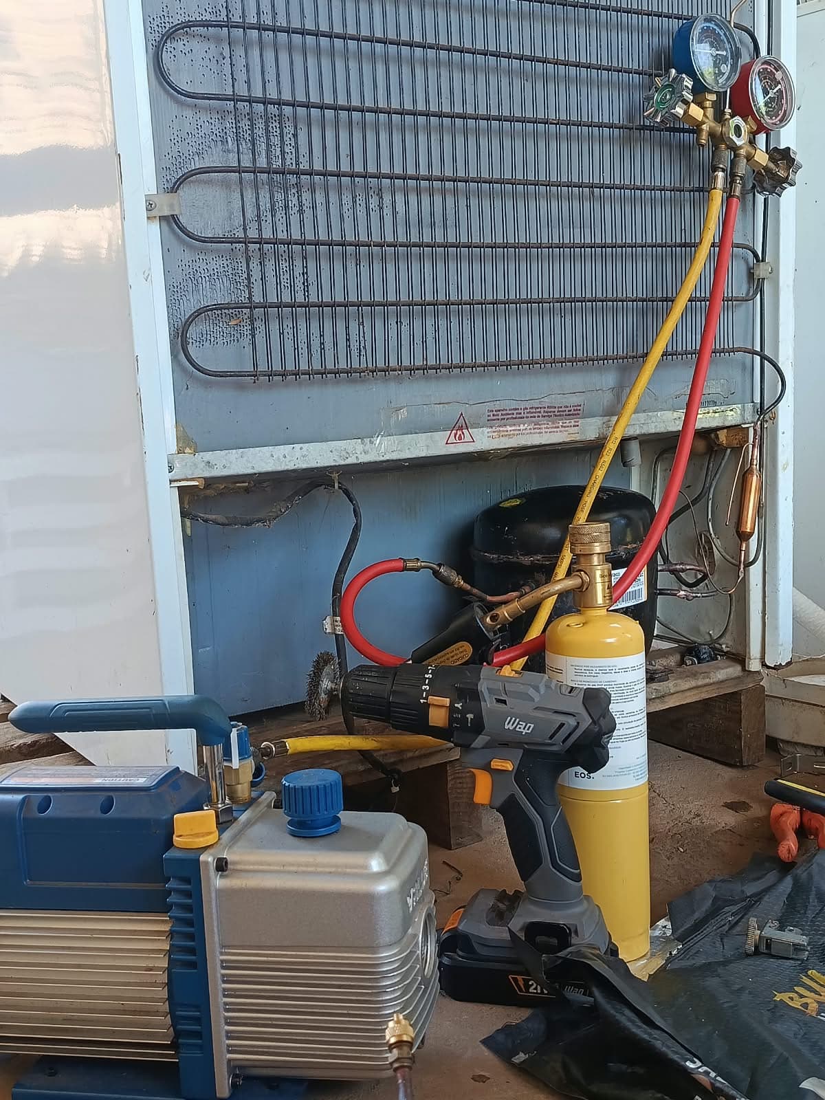 Fundo da geladeira aberto no conserto, com manifold, bomba de vácuo e cilindro de gás ligados ao compressor